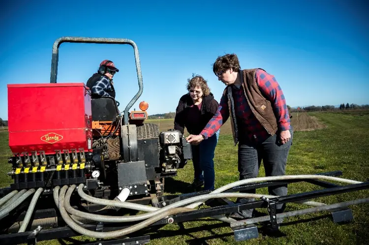 One person operates agricultural machinery while two others examine and adjust components of the attached equipment.