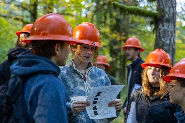 A group of students wearing orange safety helmets gathered outdoors in a forest. 